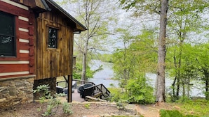 Eine Holzhütte mit Steinunterbau, einer Holzdiele und Blick auf einen von Bäumen umgebenen See.