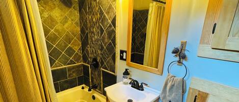 Une salle de bain avec une baignoire, un lavabo et un miroir. On y trouve un rideau de douche, un porte-serviettes et un meuble en bois.