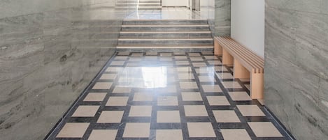 Um corredor moderno com piso xadrez, paredes de mármore e escada que leva a um andar superior.