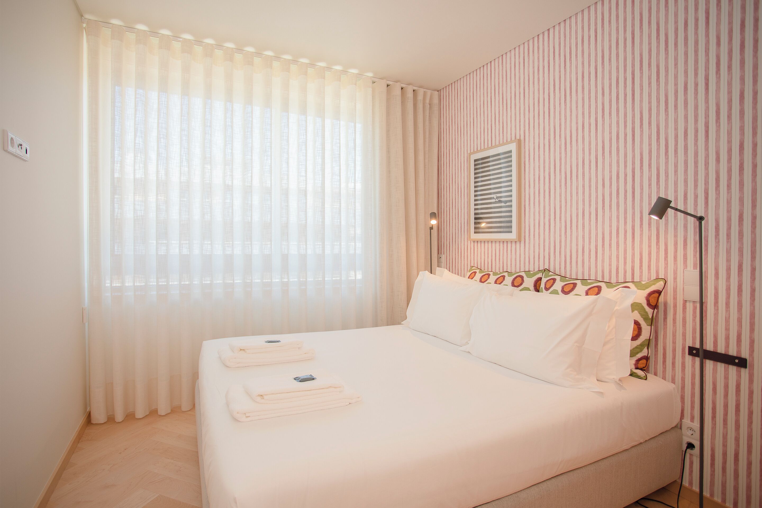 Ein ordentlich bezogenes Bett mit weißen Leinen und Handtüchern, ein Fenster mit transparenten Vorhängen und eine gestreifte Wand.