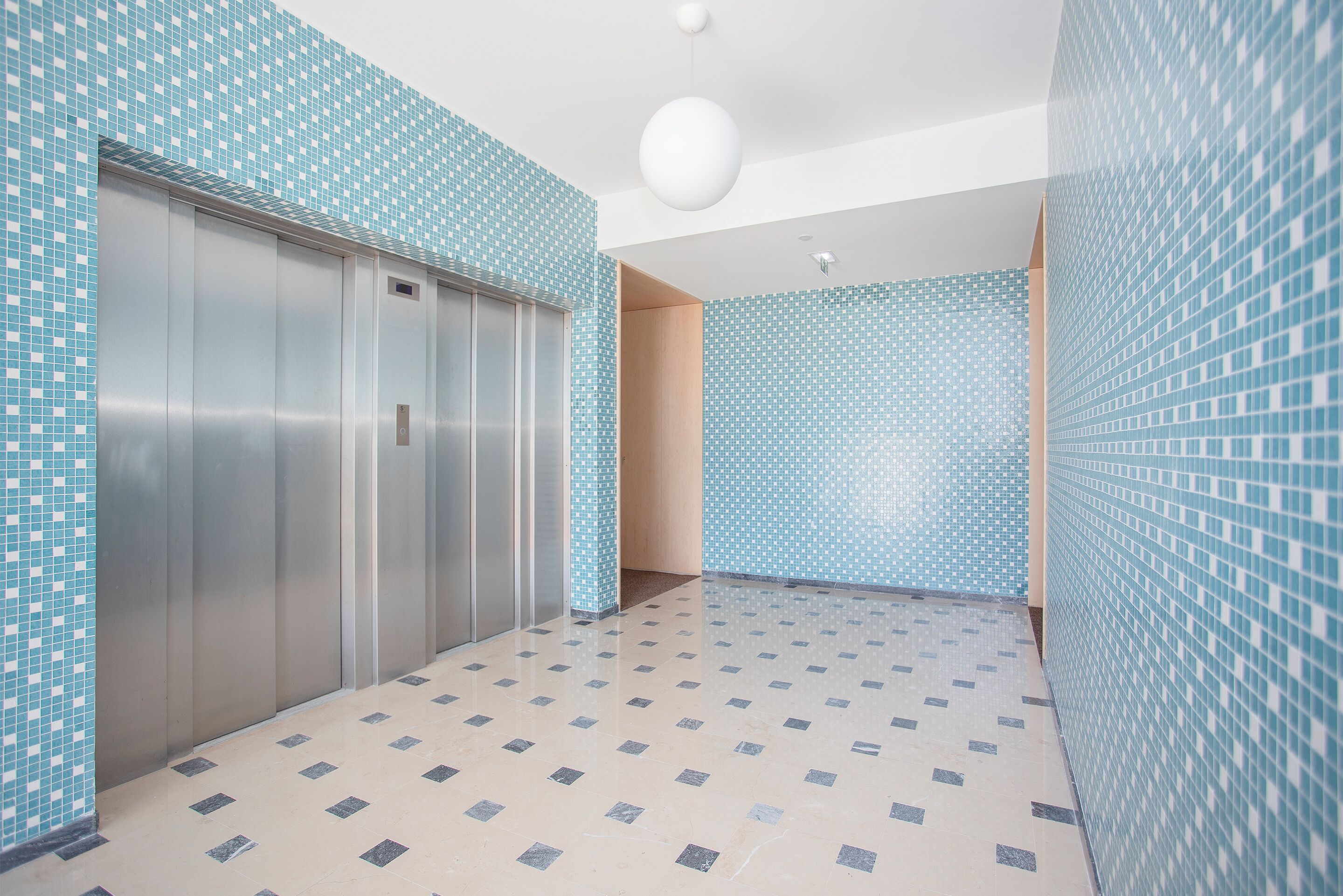 Un pasillo con suelo de baldosas, un ascensor metálico y paredes con un diseño de azulejos azules.