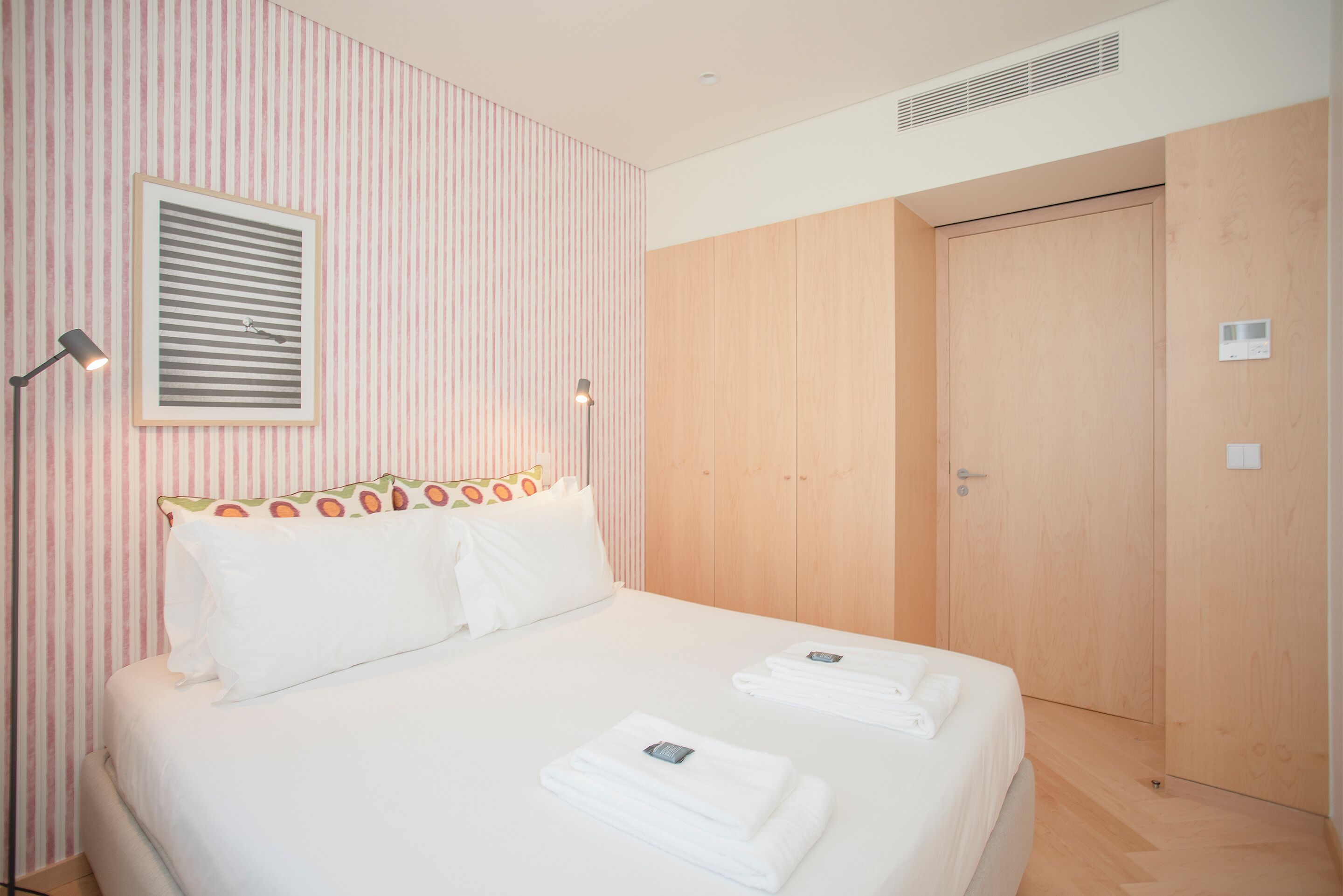 A neatly made bed with white linens, a framed picture on the wall, and a wooden wardrobe.