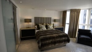 A modern hotel room with a large bed, two bedside tables with lamps, a chair, and a view of the city through large windows. - Spacius 3-bed Apartment in Kensington, London (London)
