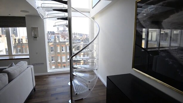 A modern living room with a spiral staircase, a large window with a city view, and a black-framed painting. - Spacius 3-bed Apartment in Kensington, London (London)
