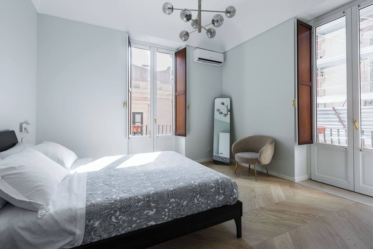 A modern bedroom with a bed, a chair, a mirror, and large windows.