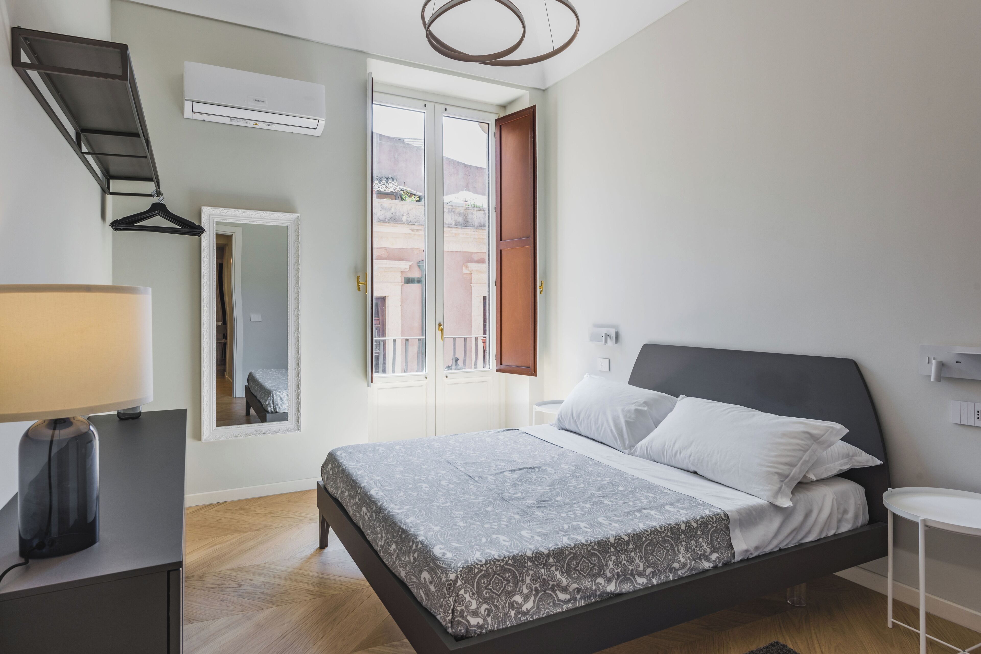 A bedroom with a bed, bedside table, lamp, mirror, and an air conditioning unit.