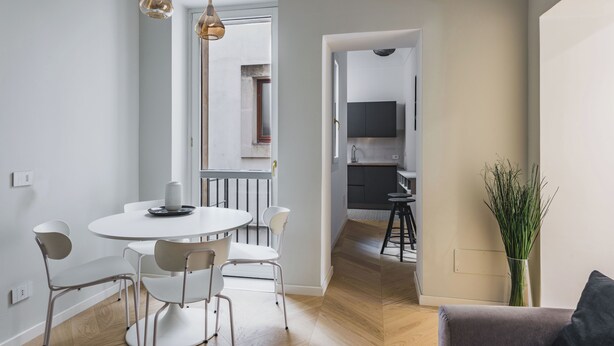 A modern dining area with a round table and chairs, a kitchen in the background, and a plant on the right. - Palazzo Bozzanca by Wonderful Italy (Syracuse)