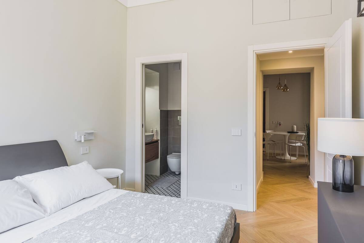 A bedroom with a bed, a bedside table, a nightstand, and a door leading to a bathroom and a dining area.