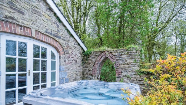 A stone building with a glass door, a brick archway, and a hot tub in the foreground. - The Stables - 2 Bedroom Cottage - Princes Gate (Narberth)
