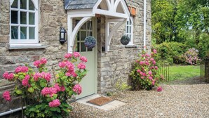 A stone house with a white door and windows, surrounded by gravel and pink hydrangea bushes. - The Stables - 2 Bedroom Cottage - Princes Gate (Narberth)