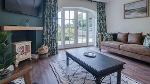 A living room with a dark wooden coffee table, a brown leather sofa, a television mounted on a dark wall, and a large window with curtains. - The Stables - 2 Bedroom Cottage - Princes Gate (Narberth)