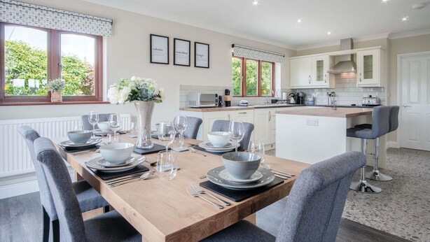 A modern kitchen with white cabinets, a central island, and a dining area with grey chairs and a wooden table set for four. - Springfield Lodge - 3 Bedroom Home - Princes Gate (Narberth)