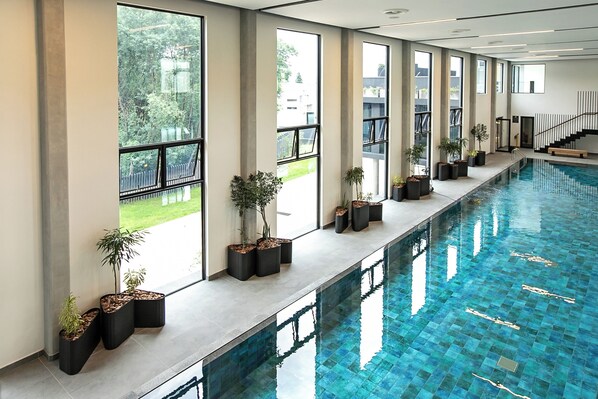 A long indoor swimming pool with large windows and potted plants along the sides.