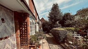 A narrow residential pathway with a brick house on the left, a garden with potted plants, and a wooden fence on the right. - Scallop Cottage Oasis of Calm - Pass the Keys (Rye)