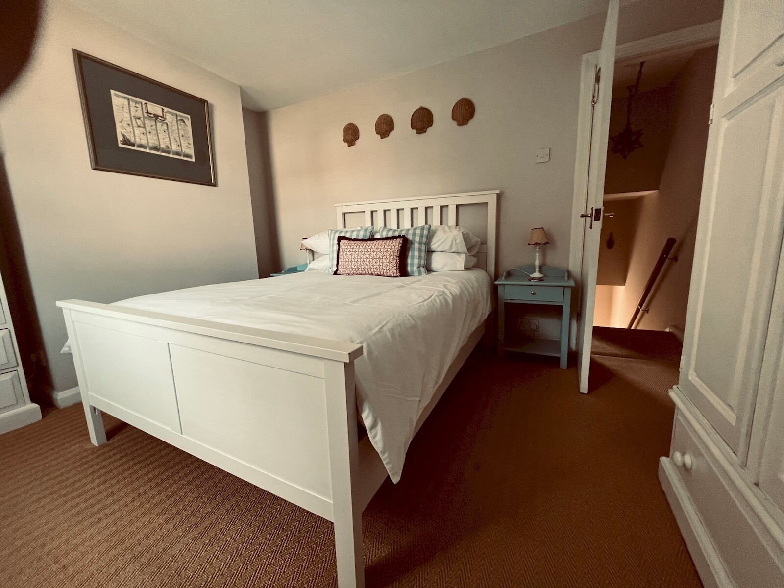 A bedroom with a white bed, a nightstand with a lamp, a framed picture on the wall, and a staircase leading to another room.