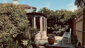 A wooden shed with a glass door, surrounded by dense foliage. A white metal chair and a large potted plant are visible in the foreground. - Scallop Cottage Oasis of Calm - Pass the Keys (Rye)