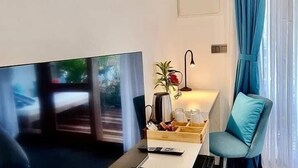 Una habitación de hotel moderna con una televisión de pantalla plana, una mesita de noche con lámpara, una planta en maceta y vistas al exterior.