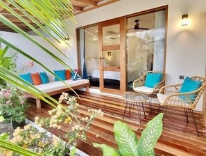 Un área de estar al aire libre acogedora con una terraza de madera, sillas de mimbre y un banco acolchado con cojines de colores.