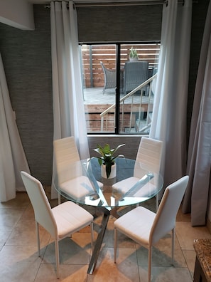 A modern dining area with a glass table and white chairs, a potted plant, and a view of an outdoor deck through a large window.