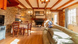 A cozy room with a stone fireplace, wooden beams, a dining table with chairs, a sofa, and a television.