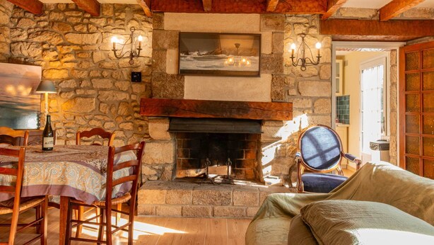 A stone fireplace with a framed picture above, wooden beams, a dining table with chairs, and a sofa.