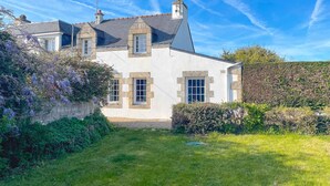 A two-story stone house with a sloped roof, multiple chimneys, and a well-maintained garden with flowering plants.