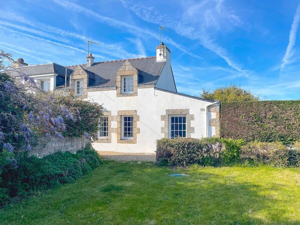 Une maison en pierre à deux étages, dotée d’un toit en pente, de plusieurs cheminées et d’un jardin bien entretenu où poussent des plantes à fleurs.