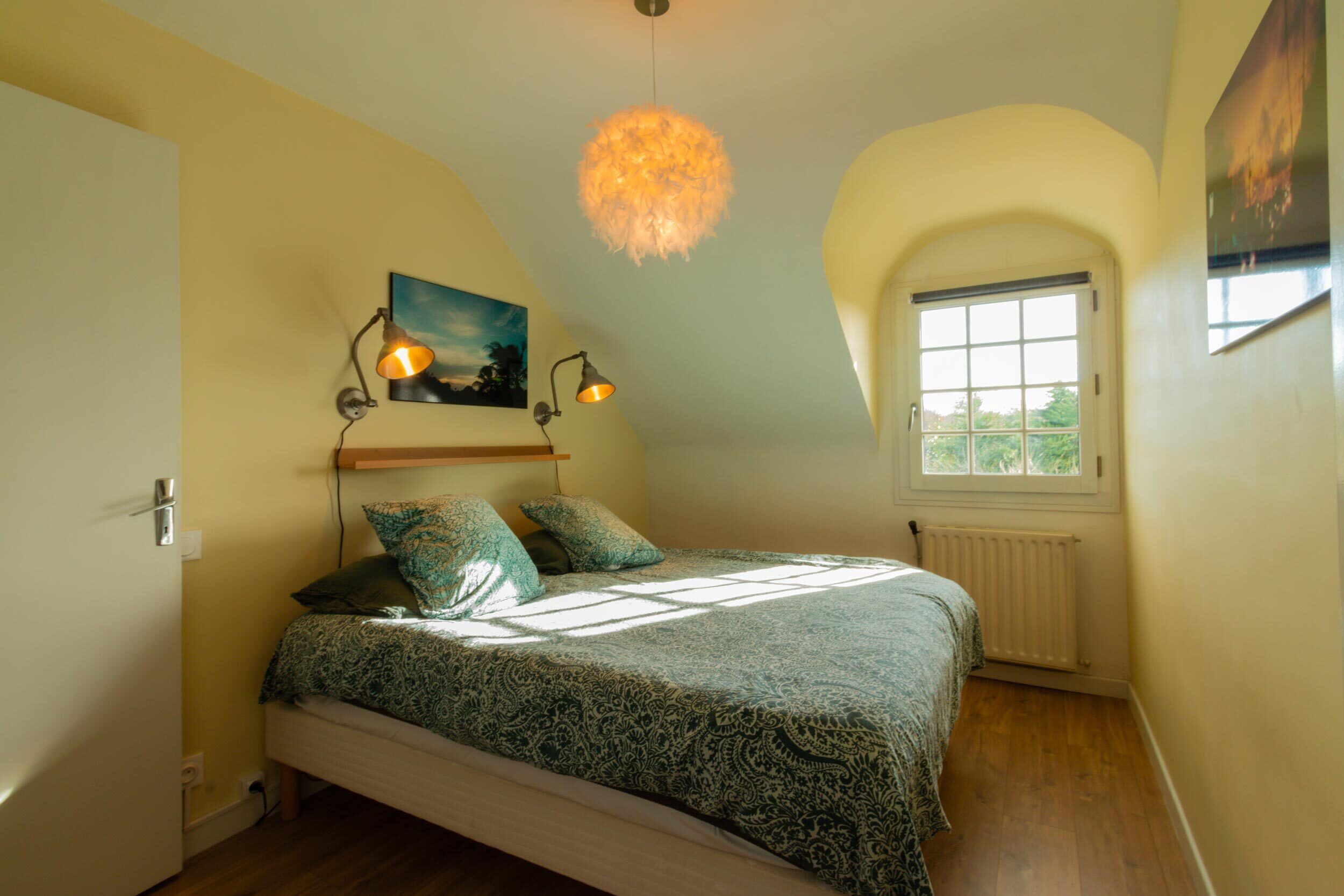 Une chambre à coucher avec un lit, deux lampes fixées au mur, un tableau, une fenêtre et un radiateur.
