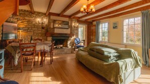 A cozy room with a stone fireplace, wooden beams, a dining area, and a large window with curtains.