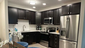 Cocina moderna con armarios oscuros, electrodomésticos de acero inoxidable y una mesa de comedor de madera con sillas azules.