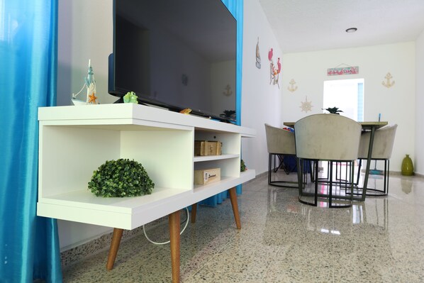 A modern living room with a flat-screen TV, a white entertainment unit, and a dining area with a table and chairs. - Unwind relax enjoy: 3br with beaches near by! (Sabana)