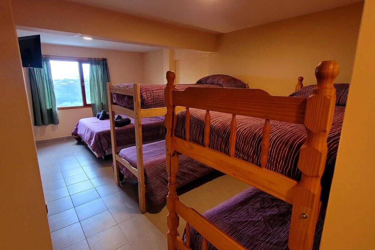 A dormitory room with three bunk beds, a television on the wall, and a window with curtains.