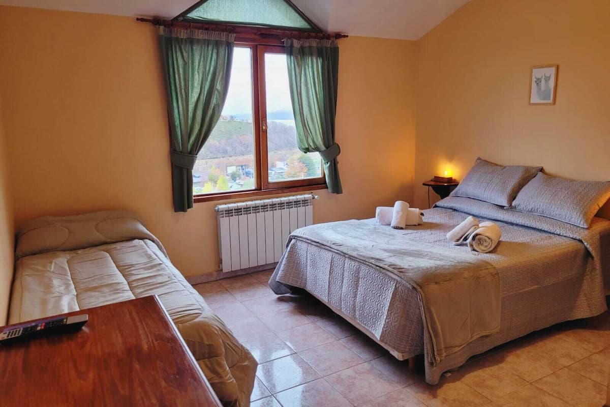A bedroom with two beds, a window with curtains, a bedside table with a lamp, and a view of the outside.