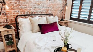 Una cama bien hecha con ropa de cama blanca y una almohada morada, una mesita de noche con lámpara, y una bandeja con alimentos para el desayuno, incluyendo cereal, fruta y una bebida.