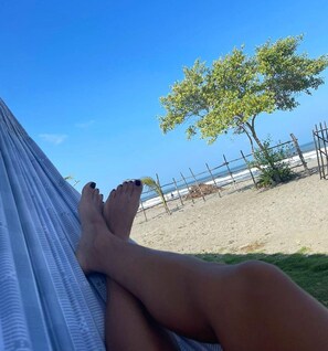 Un hamac sur lequel reposent les jambes d’une personne, avec vue sur une plage, une clôture et un arbre.