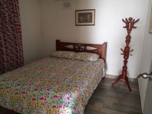 Une chambre à coucher avec une couvre-lit à motifs floraux, une tête de lit en bois, un porte-manteau en bois et une porte dotée d’une poignée argentée.