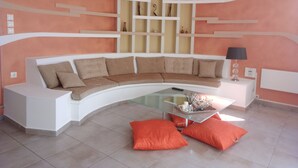 Un salon moderne avec un canapé blanc aux lignes courbes, des coussins orange, une table basse en verre et un mur doté d’étagères intégrées.