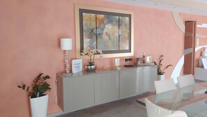 Un espace d’accueil moderne avec une table en verre, des chaises blanches, une plante en pot, un tableau abstrait encadré et un panneau indiquant : « illa Anna, un séjour élégant ».
