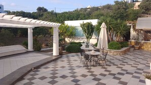 Un espace patio doté d’un sol à carreaux, d’une pergola, d’une table avec des chaises et de divers plantes en pot.