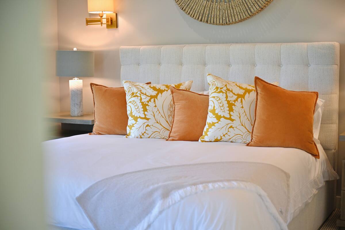A neatly made bed with white linens and patterned pillows, a tufted headboard, and a wall-mounted lamp.