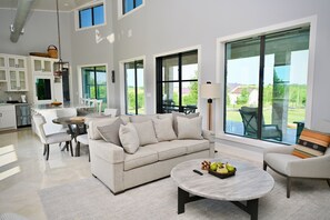 A spacious living area with a sofa, coffee table, and dining table. Large windows offer views of the outdoors.