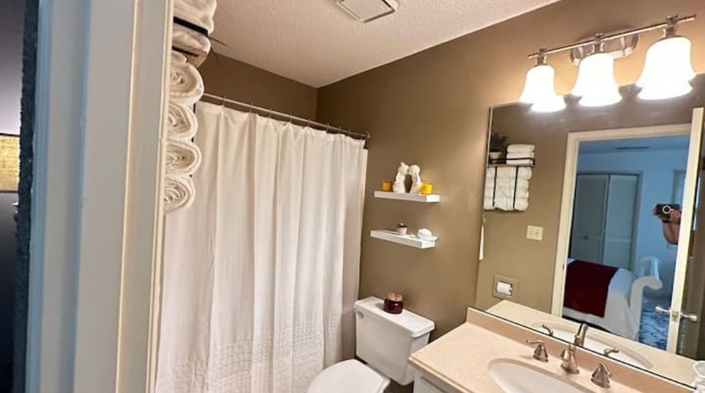A bathroom with a white shower curtain, a toilet, a sink with a mirror, and shelves with toiletries.