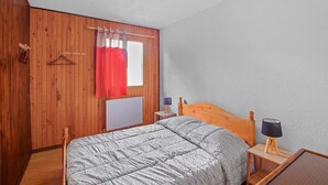 Quarto com cabeceira de madeira, cama com colcha cinza, mesa de cabeceira com abajur, janela com cortinas vermelhas e parede de madeira.