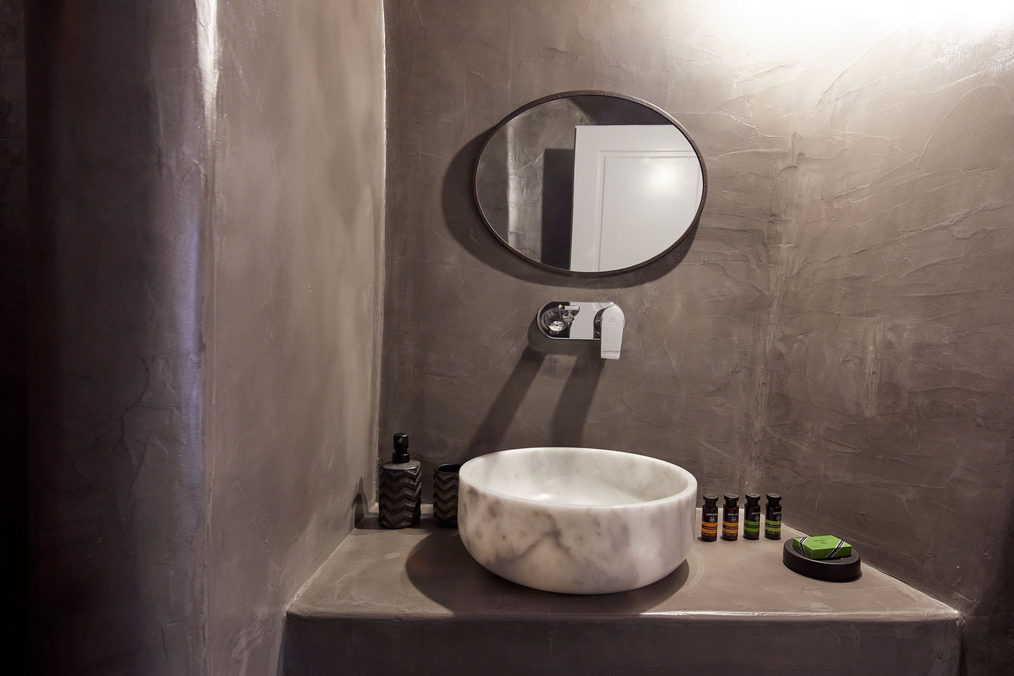 A bathroom with a marble sink, a round mirror, a towel dispenser, and a few toiletries on the counter.