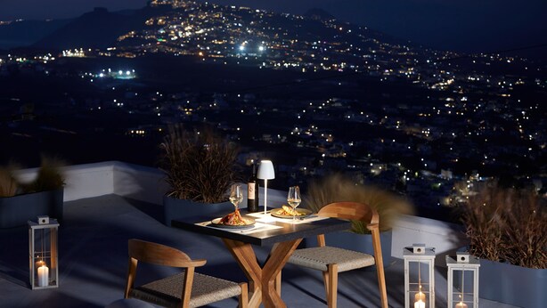 Un área de comedor al aire libre con mesa y sillas, velas, y vista a un paisaje urbano de noche.