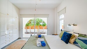 A bedroom with a bed, a bedside table, a lamp, and a view of an outdoor area with trees and a wooden fence. - The Bridge Villa by Ezoria Villas (Paphos)