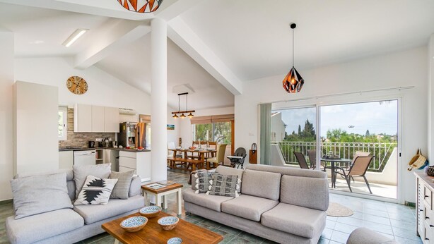 A spacious living area with a sofa set, a coffee table, and a dining table. There is a kitchen in the background and a balcony with outdoor seating. - The Bridge Villa by Ezoria Villas (Paphos)