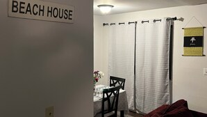 Ein Zimmer mit einem Schild, das "BEACH HOUSE" zeigt, einem Esstisch mit Stühlen, einem roten Sofa und einem Vorhang mit Muster.