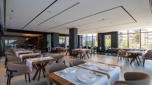 A spacious dining area with multiple tables set for dining, large windows offering views of the city, and a modern interior design. - Mango Luxury Resorts (Sarandë)