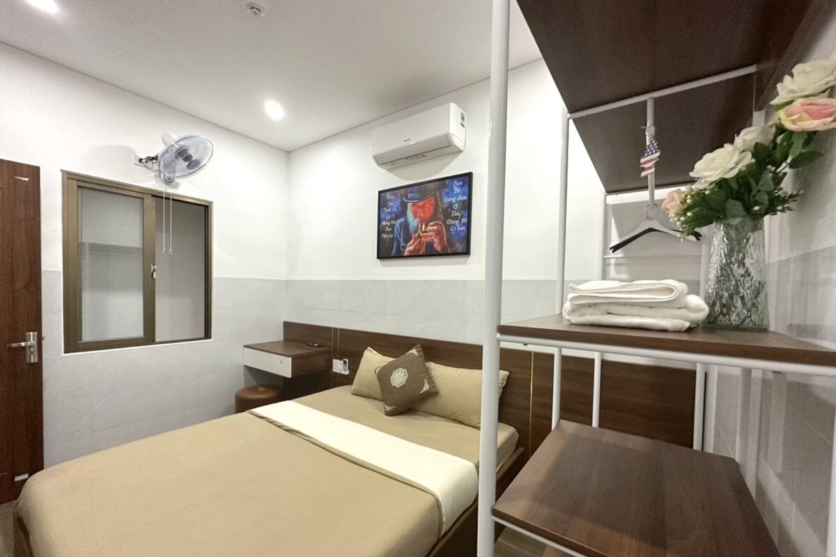 A hotel room with a bed, a bedside table, a small desk, an air conditioning unit, a wall-mounted TV, and a window with a view of the city.